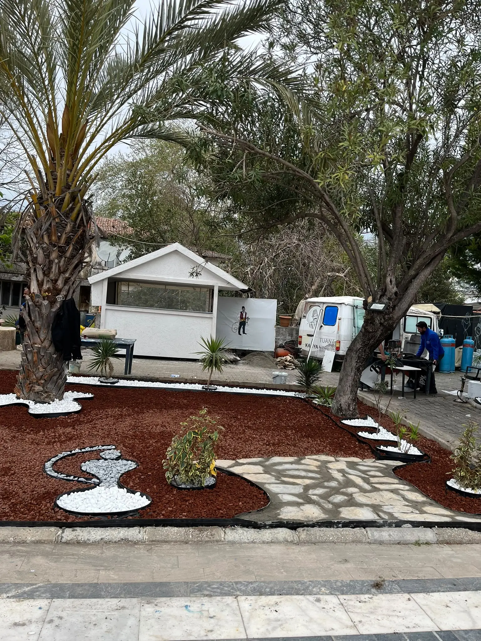 Garten mit ausgewachsenen Bäumen, rotem Mulch und hellen Steinwegen